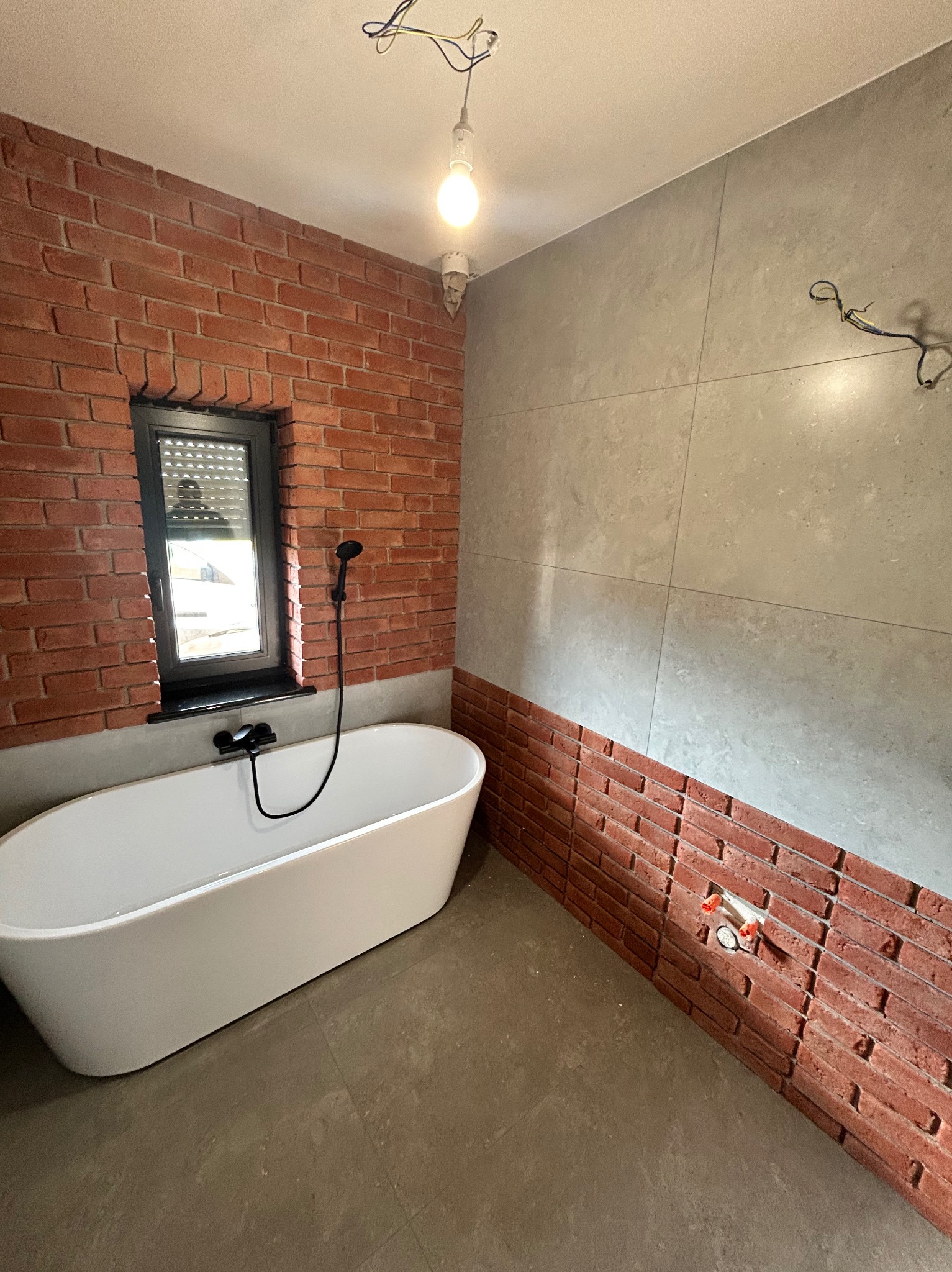 Nowoczesna łazienka z wanną wolnostojącą, ceglaną ścianą i szarymi płytkami. Widoczne okno z roletami i minimalistyczna armatura w czarnym kolorze.