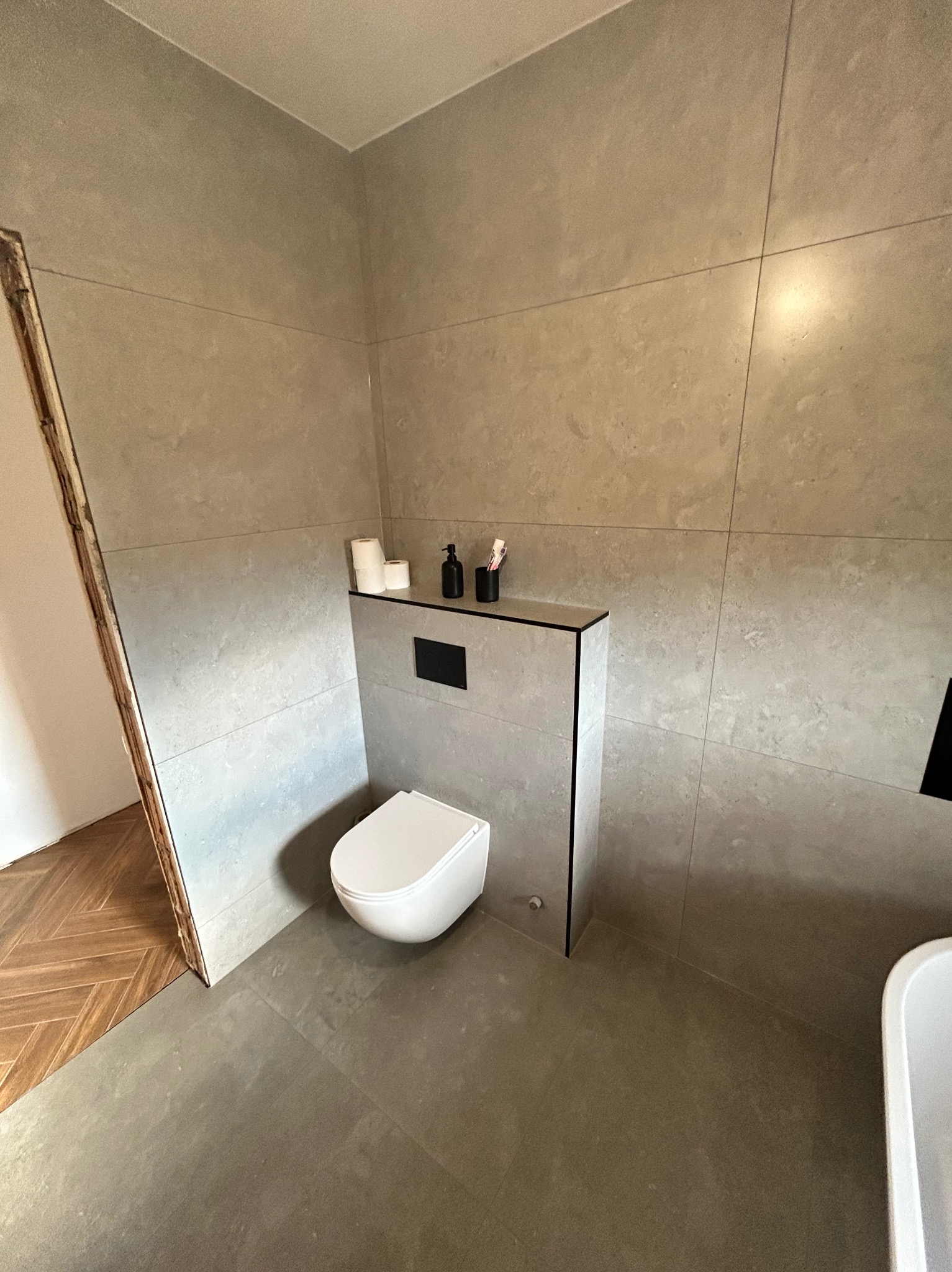 Nowoczesna łazienka z szarymi płytkami wielkoformatowymi, podwieszaną toaletą i fragmentem drewnianej podłogi w jodełkę przy wejściu. Minimalistyczny design.