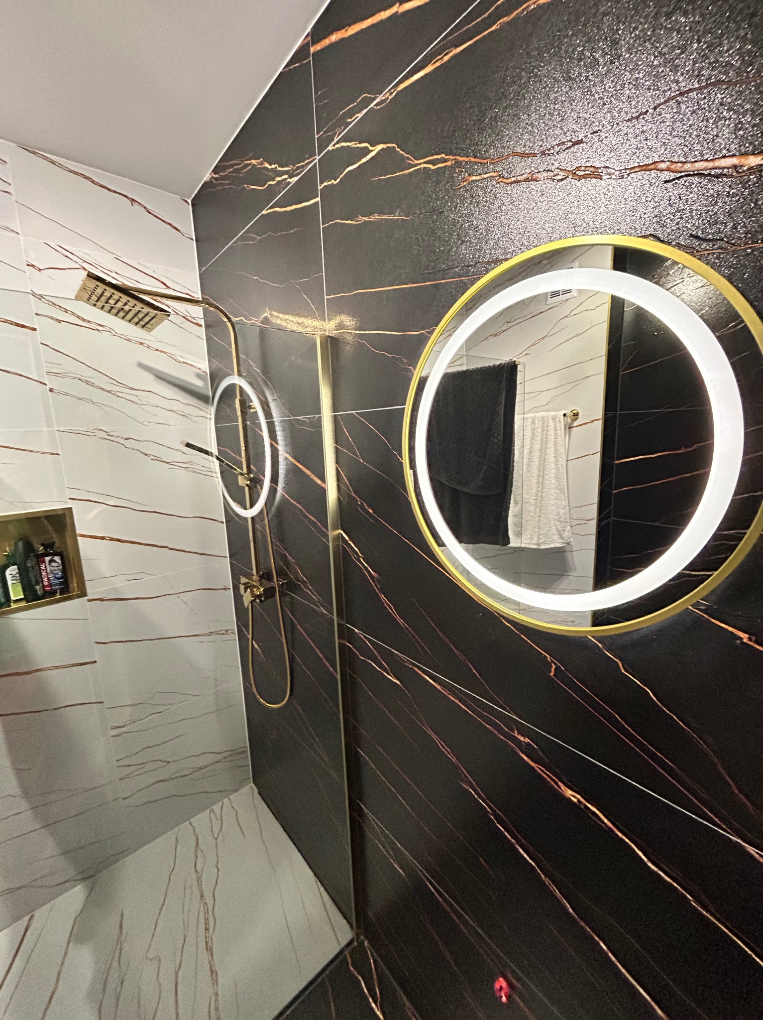 Nowoczesna kabina prysznicowa z czarnymi płytkami imitującymi marmur z złotymi żyłami, okrągłe lustro z podświetleniem LED i złotą armaturą.