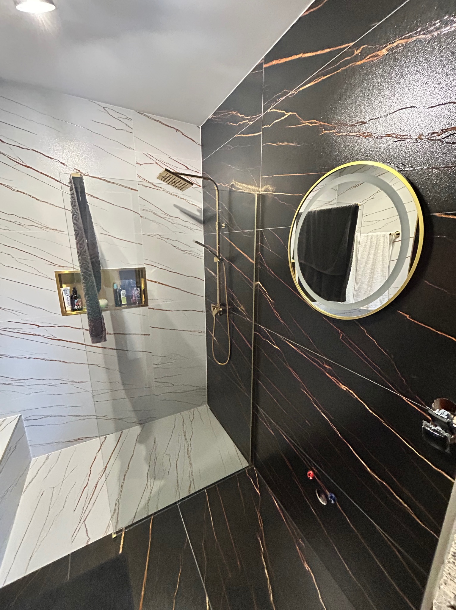 Wykończona łazienka z czarno-białymi płytkami imitującymi marmur, złotą armaturą i okrągłym lustrem z podświetleniem LED. W ścianie wnęka z kosmetykami.