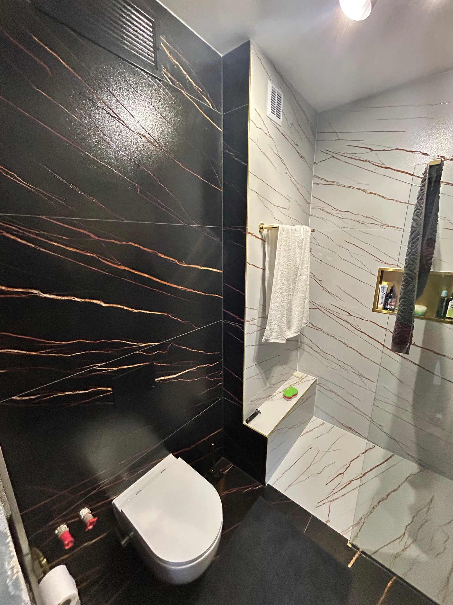 Elegancka łazienka z czarno-białymi płytkami imitującymi marmur z miedzianymi żyłami, wisząca toaleta, złote akcenty i wnęka prysznicowa.