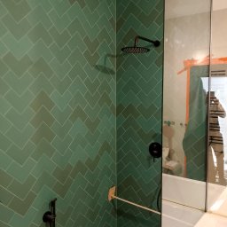 Kraina Wykończeń Daniel Witczak Łódź - Wykończona łazienka z zielonymi płytkami ułożonymi w jodełkę, czarną armaturą i szklaną kabiną prysznicową. Widoczne odbicie osoby robiącej zdjęcie.