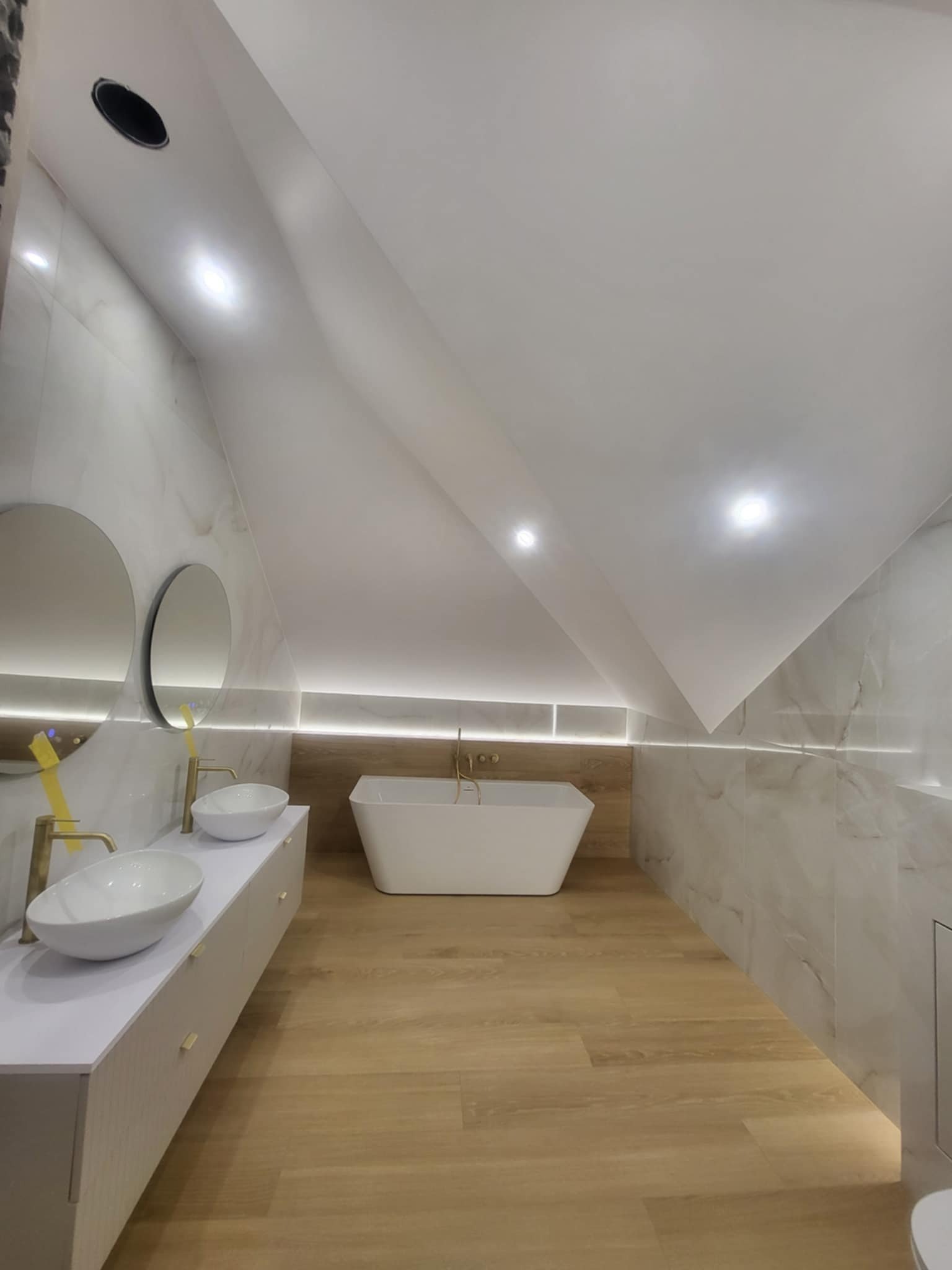 Nowoczesna łazienka na poddaszu z marmurowymi płytkami, drewnianą podłogą, wolnostojącą wanną i umywalkami nablatowymi z okrągłymi lustrami.