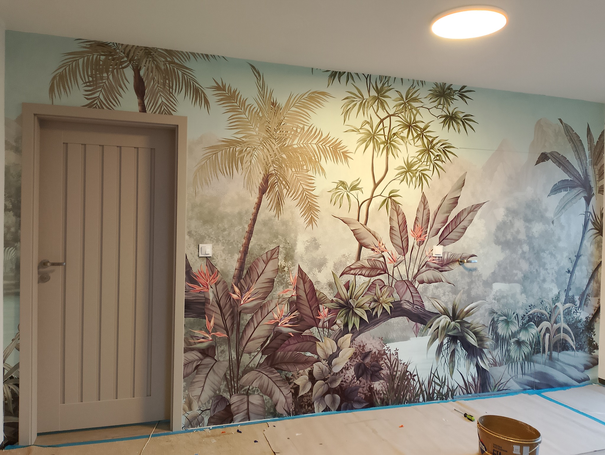 Pokój z fototapetą w motywy tropikalne: palmy, góry i roślinność. Widoczna beżowa, drewniana futryna drzwi i wiaderko z farbą na podłodze, co sugeruje świeżo zakończone prace.