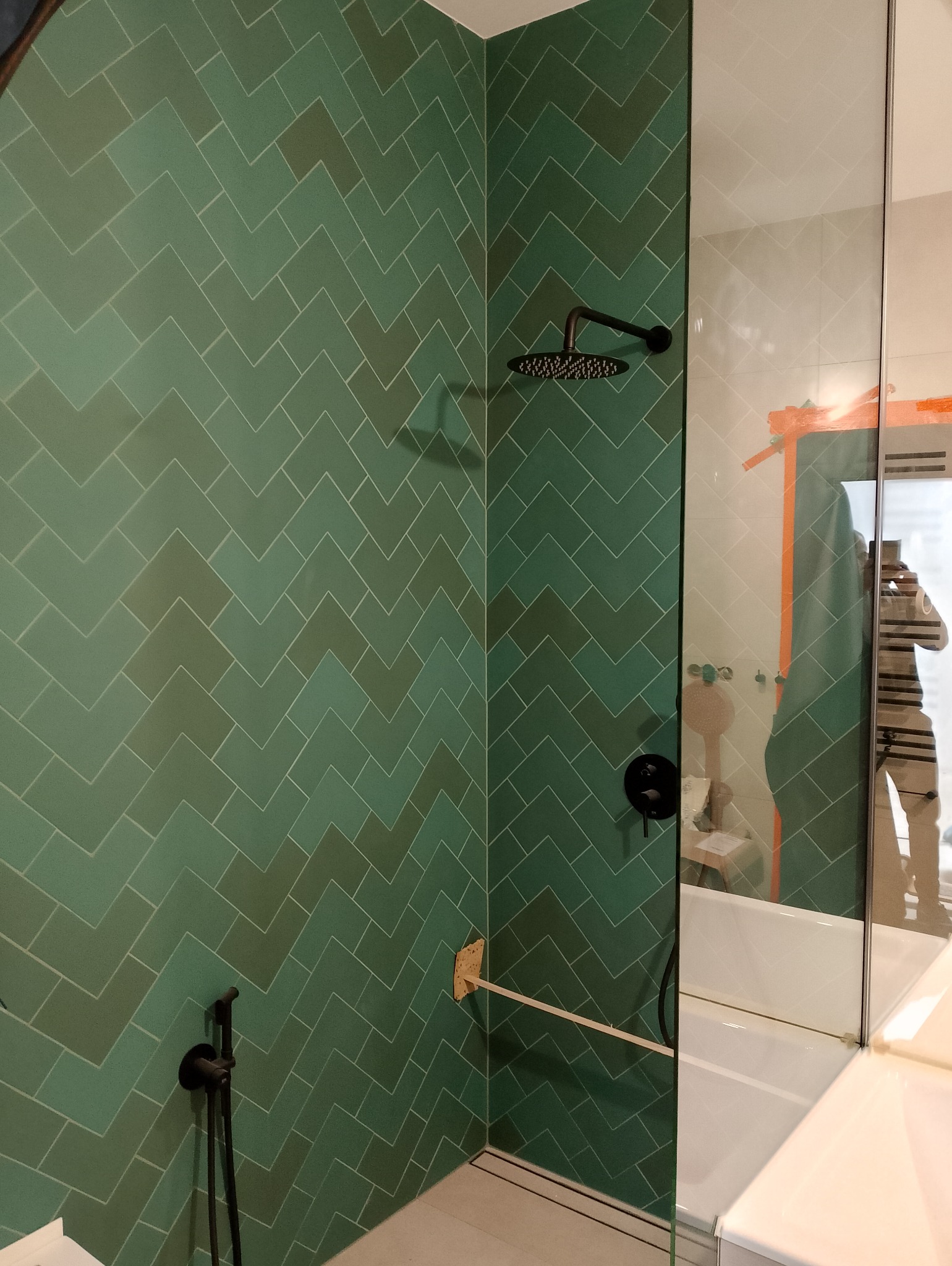 Wykończona łazienka z zielonymi płytkami ułożonymi w jodełkę, czarną armaturą i szklaną kabiną prysznicową. Widoczne odbicie osoby robiącej zdjęcie.