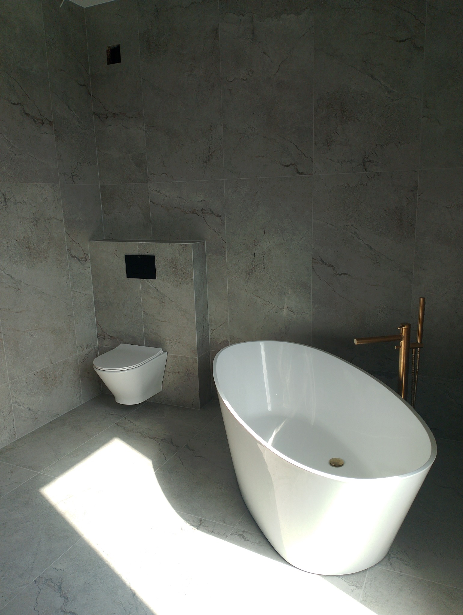 Nowoczesna łazienka z wolnostojącą wanną i podwieszaną toaletą, wykończona płytkami imitującymi marmur. Złota armatura dodaje elegancji. Wnętrze doświetlone naturalnym światłem.