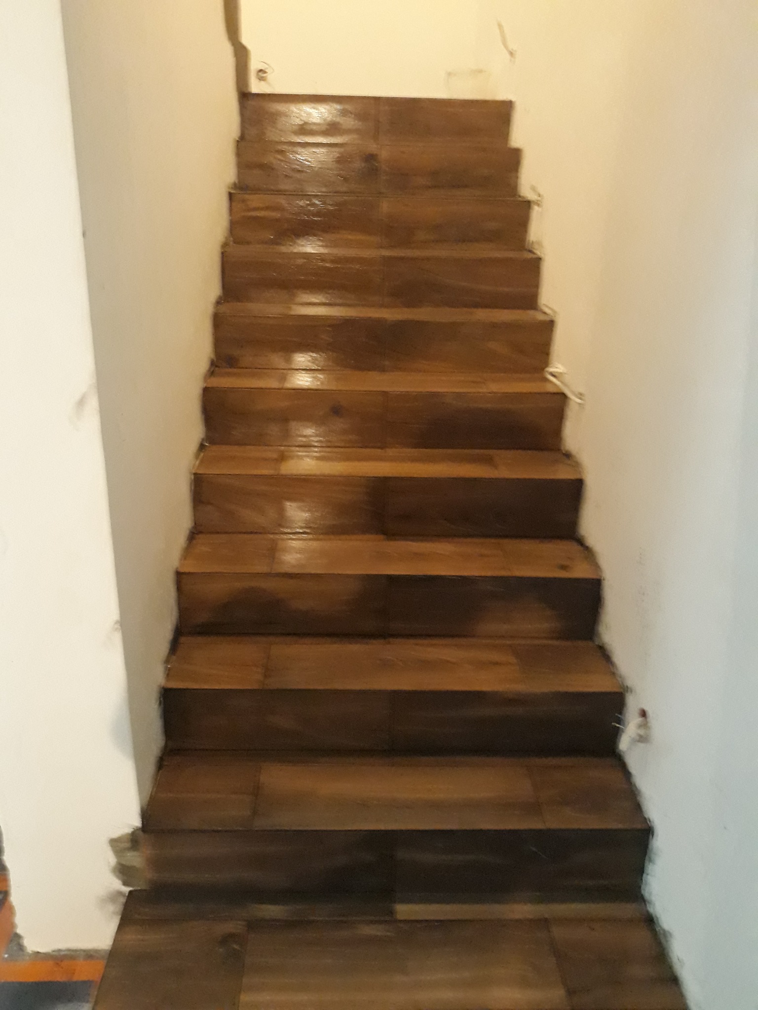 Wykończone schody z ciemnego drewna, prowadzące w górę. Widoczne niedociągnięcia przy ścianie, co sugeruje etap końcowy prac wykończeniowych. Ujęcie z dołu.