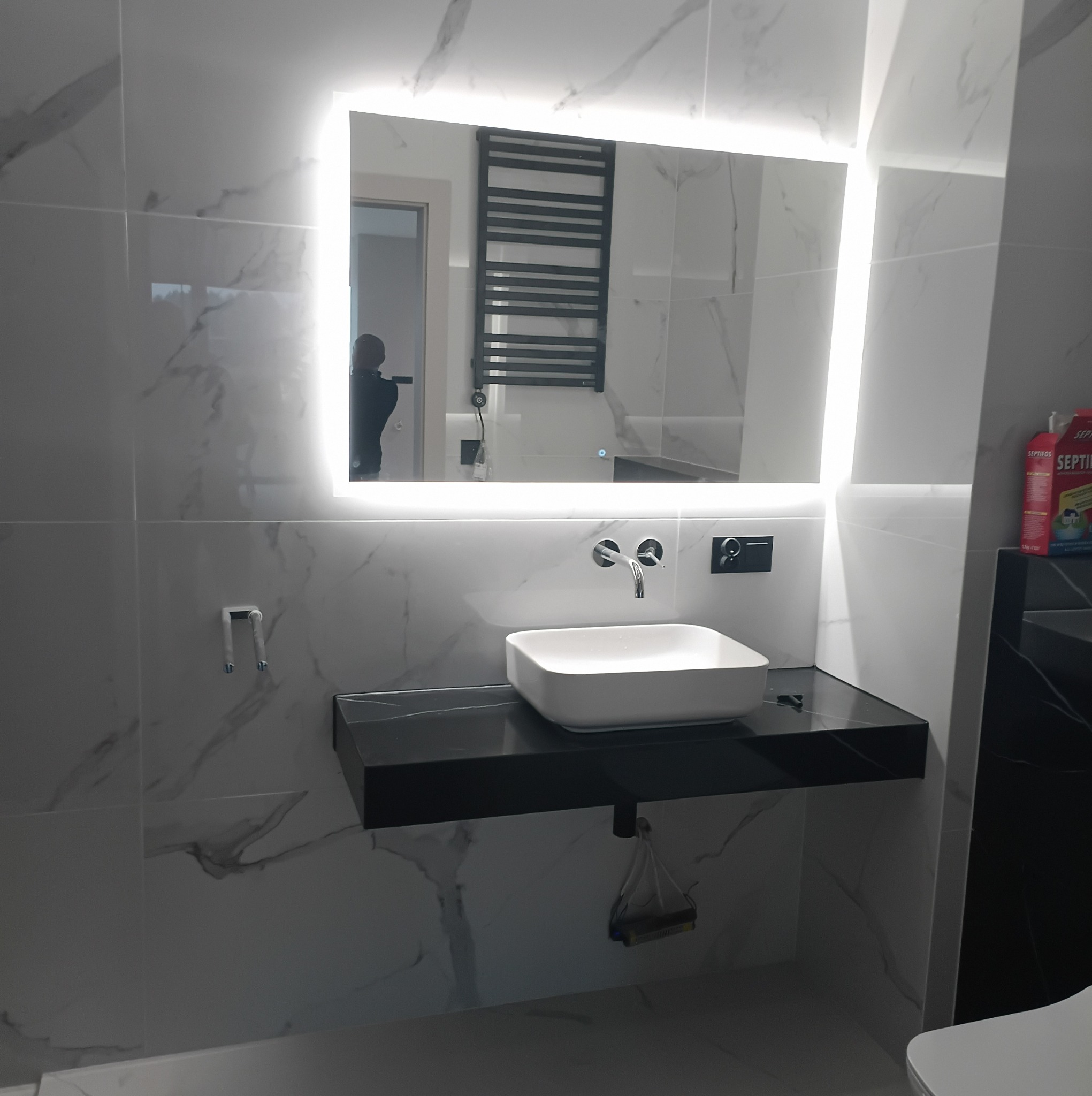 Nowoczesna łazienka z białymi płytkami imitującymi marmur, czarnym blatem pod umywalką i podświetlanym lustrem. Widoczny grzejnik drabinkowy i armatura.