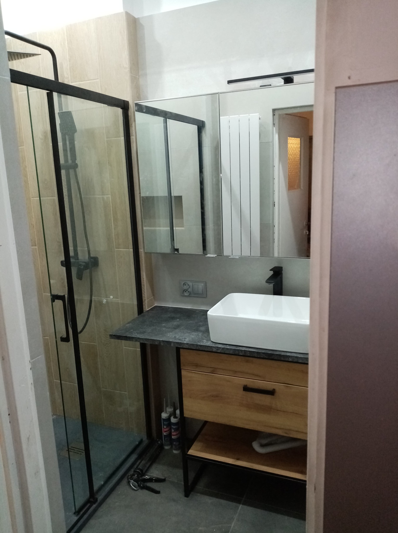 Nowoczesna łazienka z kabiną prysznicową typu walk-in, umywalką nablatową na szafce z drewnianym frontem i czarną armaturą. Widoczne płytki w odcieniach beżu i szarości.