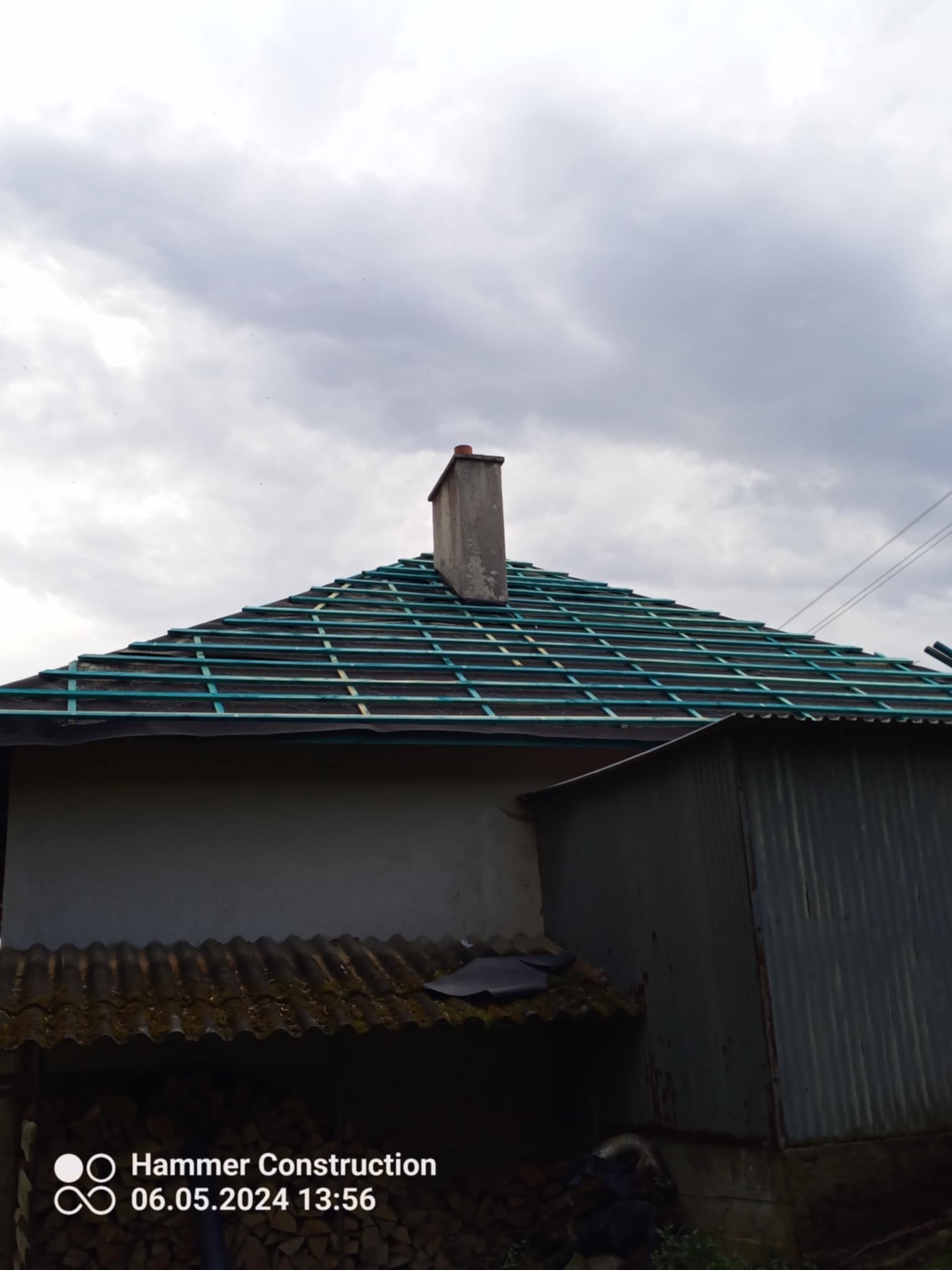 Dach z łatami pod nowe pokrycie, widoczny komin i fragment budynku. Niebo pochmurne. Etap prac dekarskich, przygotowanie do krycia dachu.