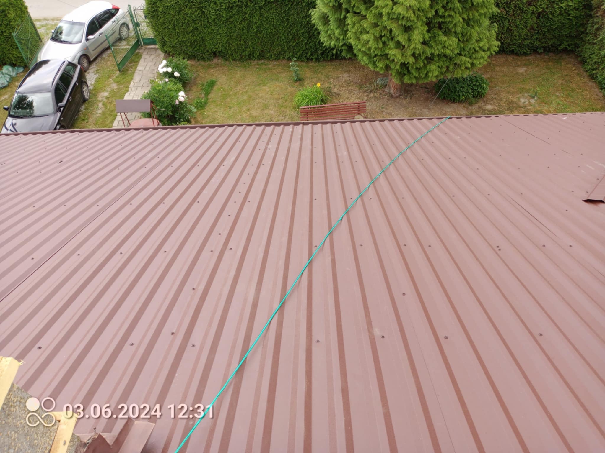Widok z góry na brązowy dach z blachy trapezowej, z zielonym przewodem biegnącym po przekątnej. W tle ogród z dwoma samochodami i zielenią.