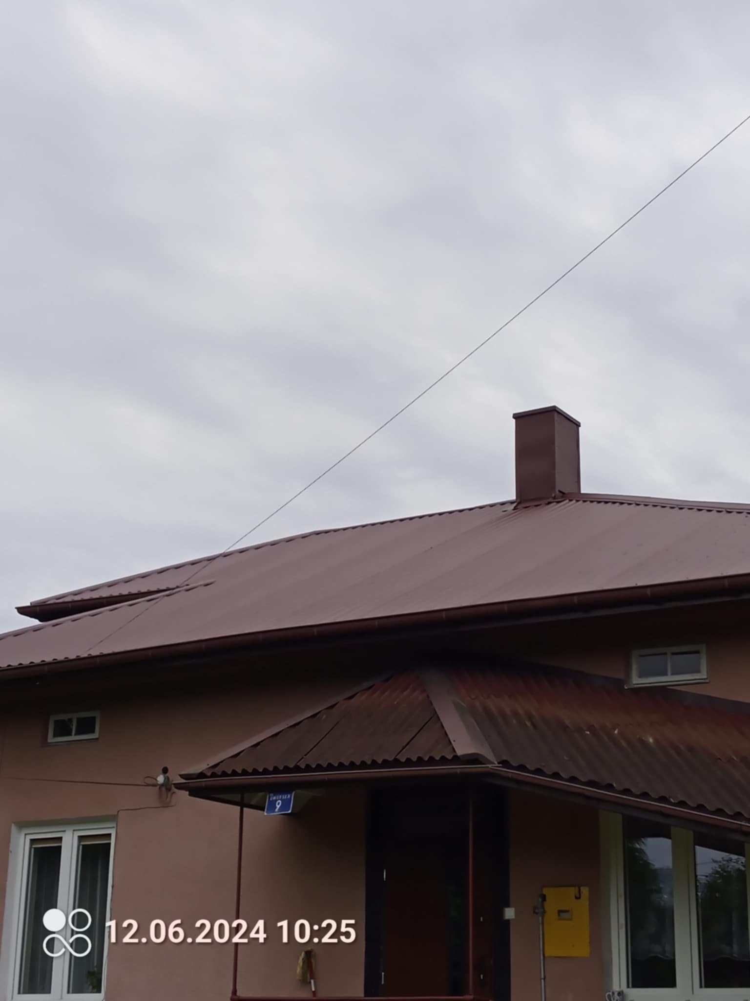 Dom z brązowym dachem z blachy trapezowej i kominem. Widoczny fragment elewacji i numer domu. Pochmurne niebo w tle.