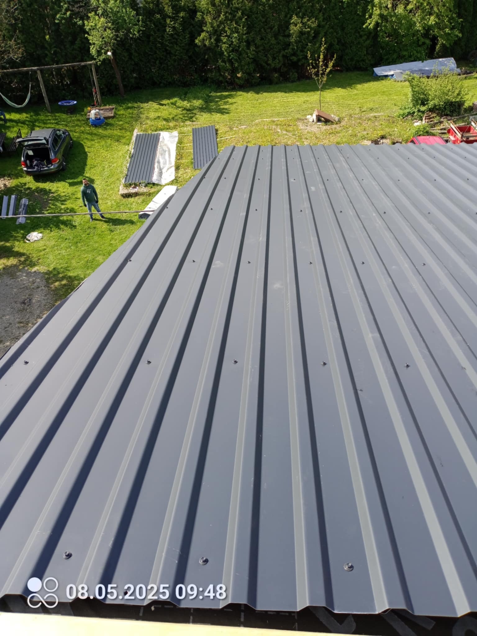 Szary dach z blachy trapezowej, widok z góry, na tle zielonego ogrodu z drzewami i samochodem. Widoczne arkusze blachy przygotowane do montażu.