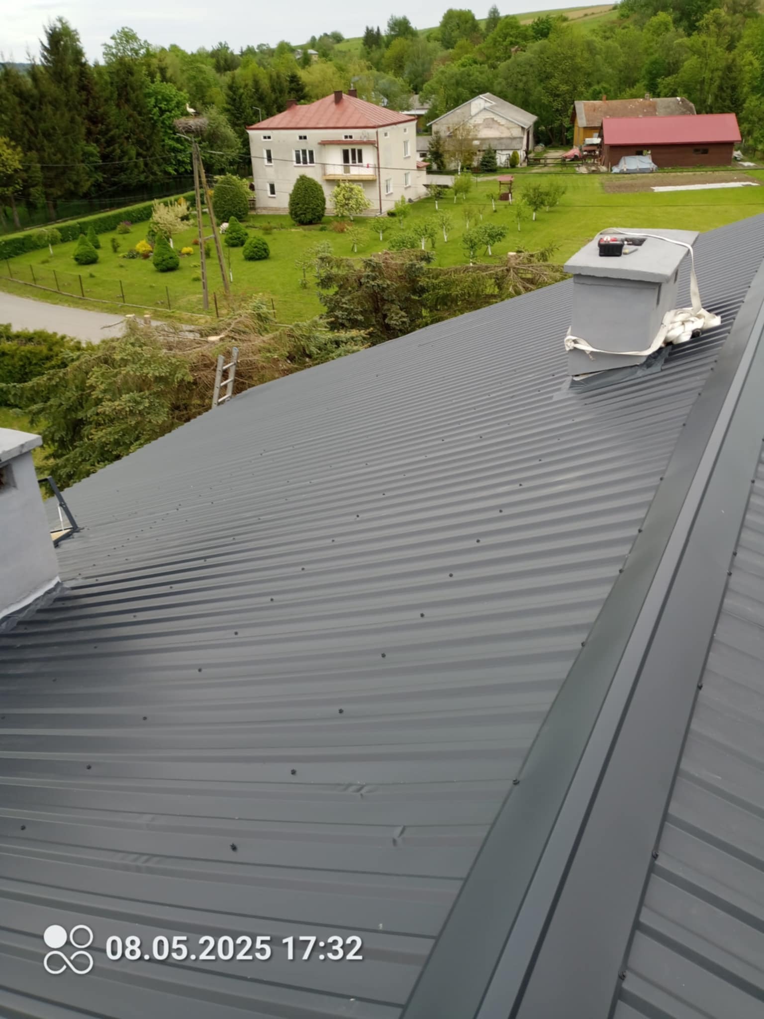 Szary dach z blachy trapezowej po montażu, widok z góry na tle zabudowań i zieleni. Kominek zabezpieczony taśmą. Widoczny fragment drabiny. Data i godzina w rogu.