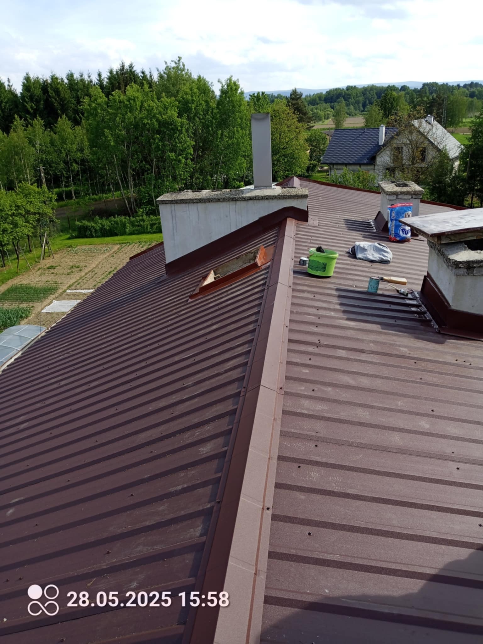 Brązowy dach z blachy trapezowej, widoczne kominy, narzędzia i akcesoria dekarskie. W tle zieleń drzew i zabudowania. Ujęcie z góry, lekko z ukosa.