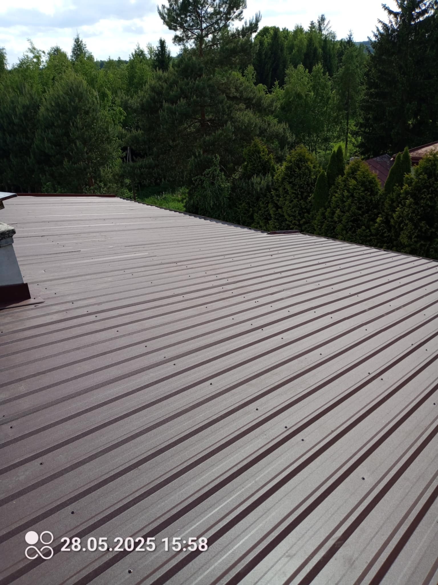 Brązowy dach z blachy trapezowej, widok z góry, na tle zieleni drzew. Widoczny komin i data wykonania zdjęcia w lewym dolnym rogu.