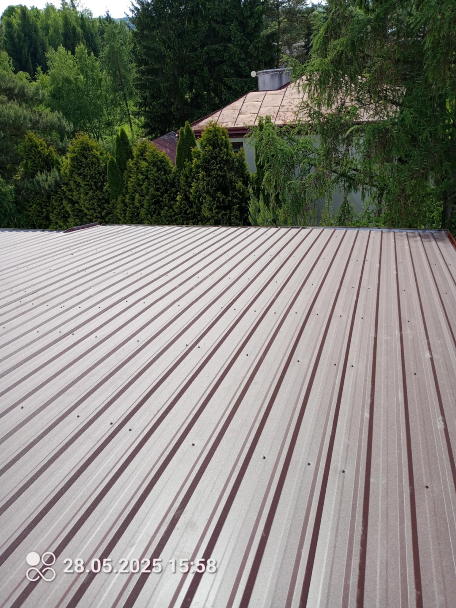 Nowy, brązowy dach z blachy trapezowej, widok z góry na tle zieleni drzew. Widoczne mocowania blachy. Data i godzina w rogu zdjęcia.