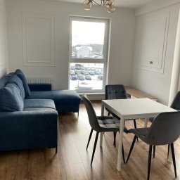 Wykończenie wnętrz Wiktor Buczyński - Jasne wnętrze: narożnik w odcieniach niebieskiego, stół z krzesłami na drewnianej podłodze. W tle okno z widokiem na parking i budynek. Minimalistyczny design.