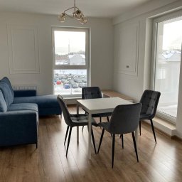 Wykończenie wnętrz Wiktor Buczyński - Jasne wnętrze salonu z niebieską sofą narożną, białym stołem i szarymi krzesłami. Widok na parking przez okno. Parkiet na podłodze.