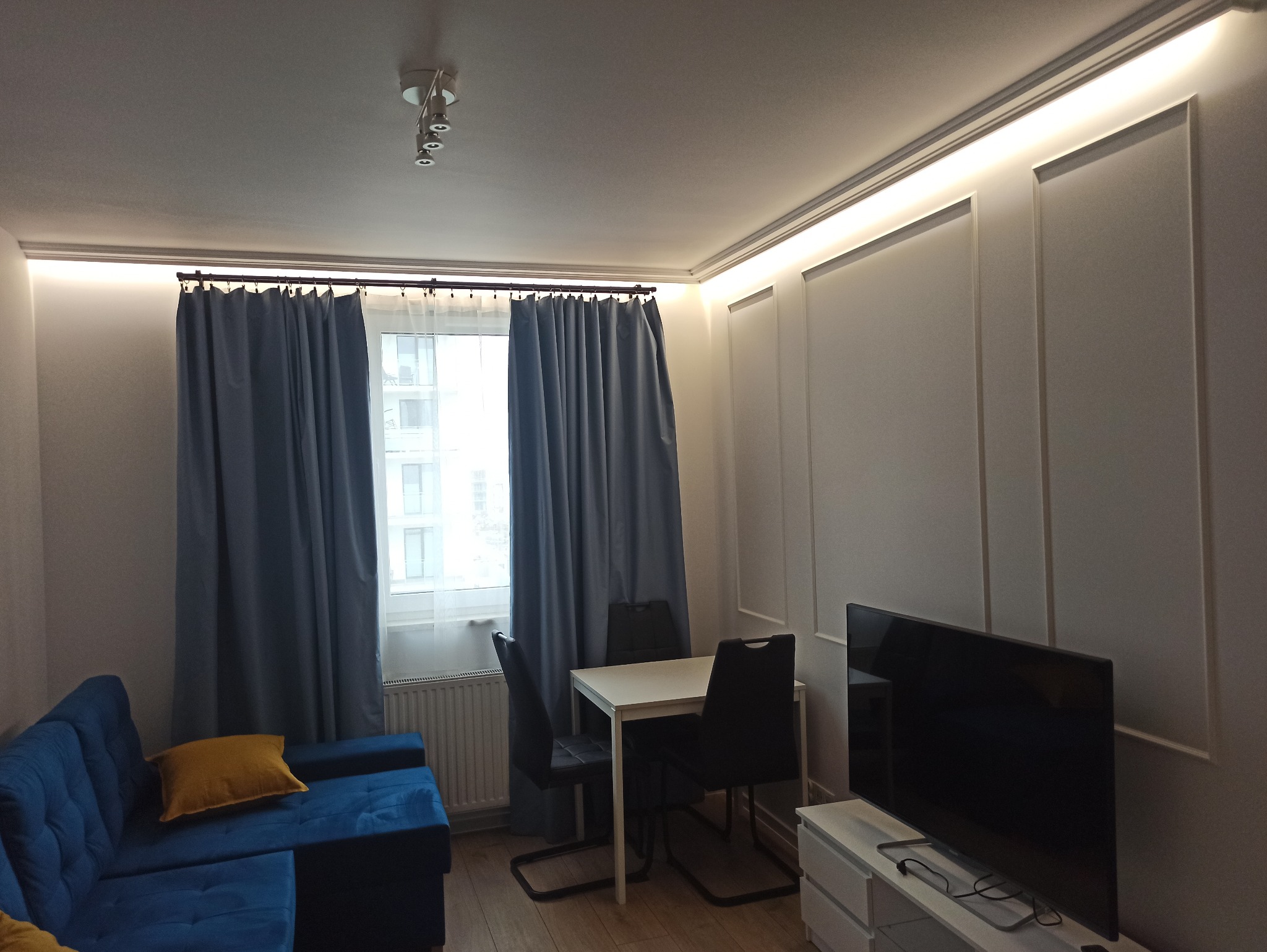 Nowoczesny salon z niebieską sofą, stołem i telewizorem. Ściany zdobią panele dekoracyjne z oświetleniem LED. Okno zasłonięte niebieskimi zasłonami.