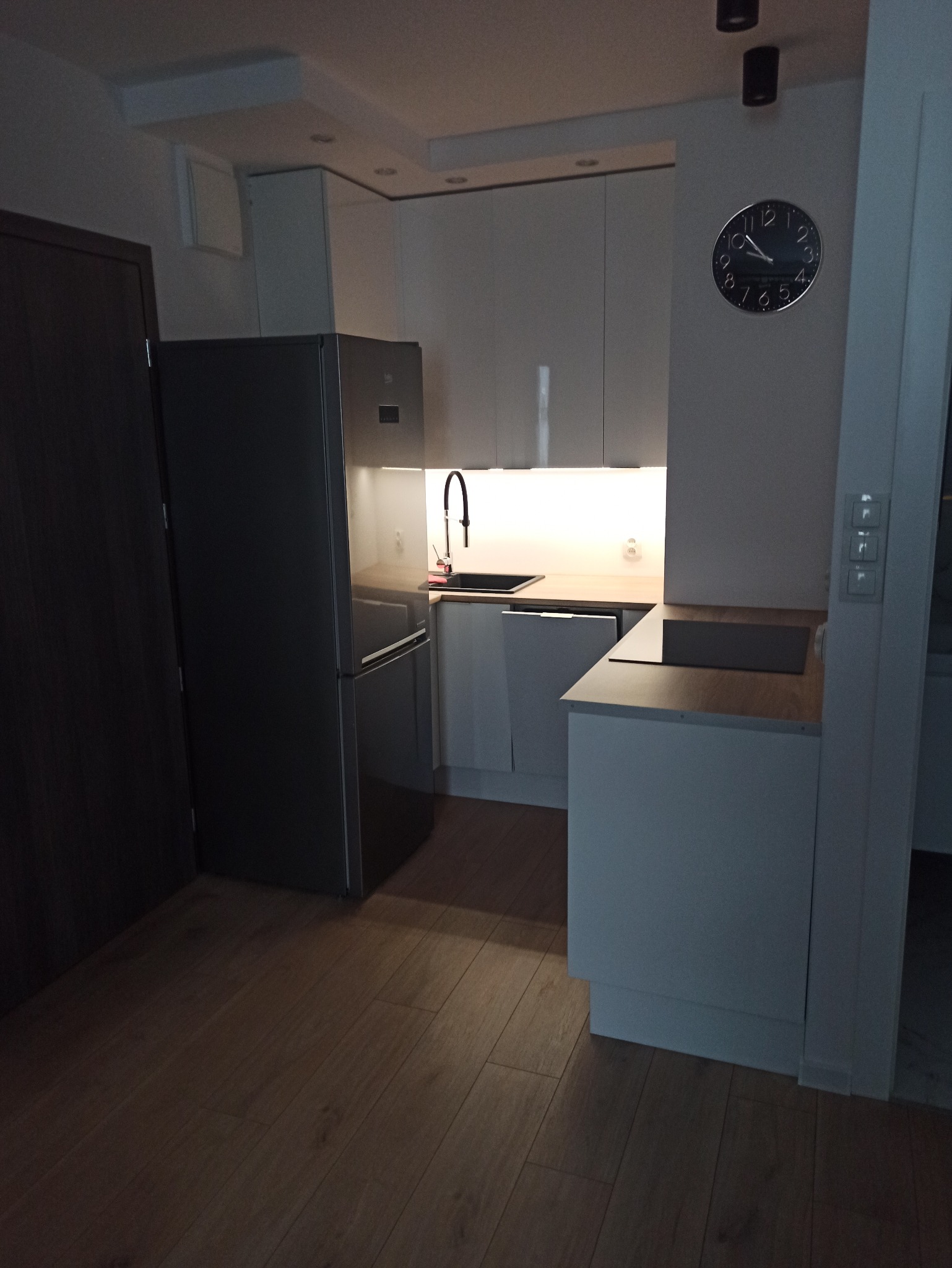 Nowoczesna, minimalistyczna kuchnia z białymi szafkami, drewnianym blatem i srebrną lodówką. Na ścianie okrągły zegar. Podłoga z paneli.