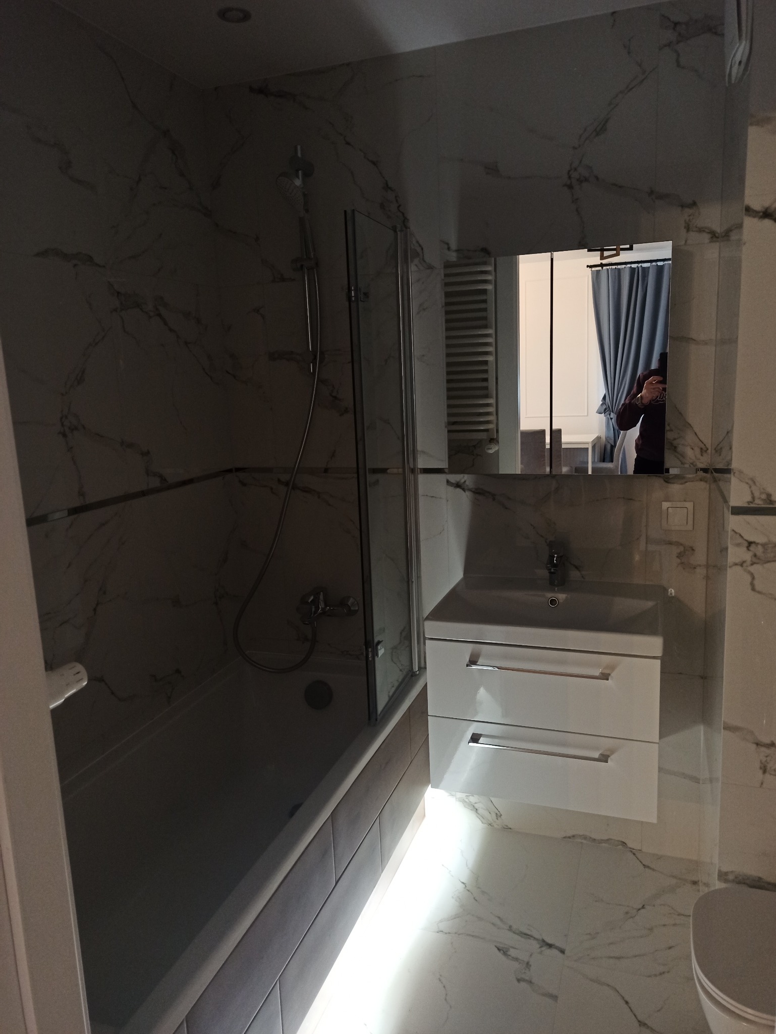 Biała łazienka z marmurowymi płytkami, kabiną prysznicową i szafką pod umywalką. W lustrze odbija się fragment innego pomieszczenia z zasłonami.