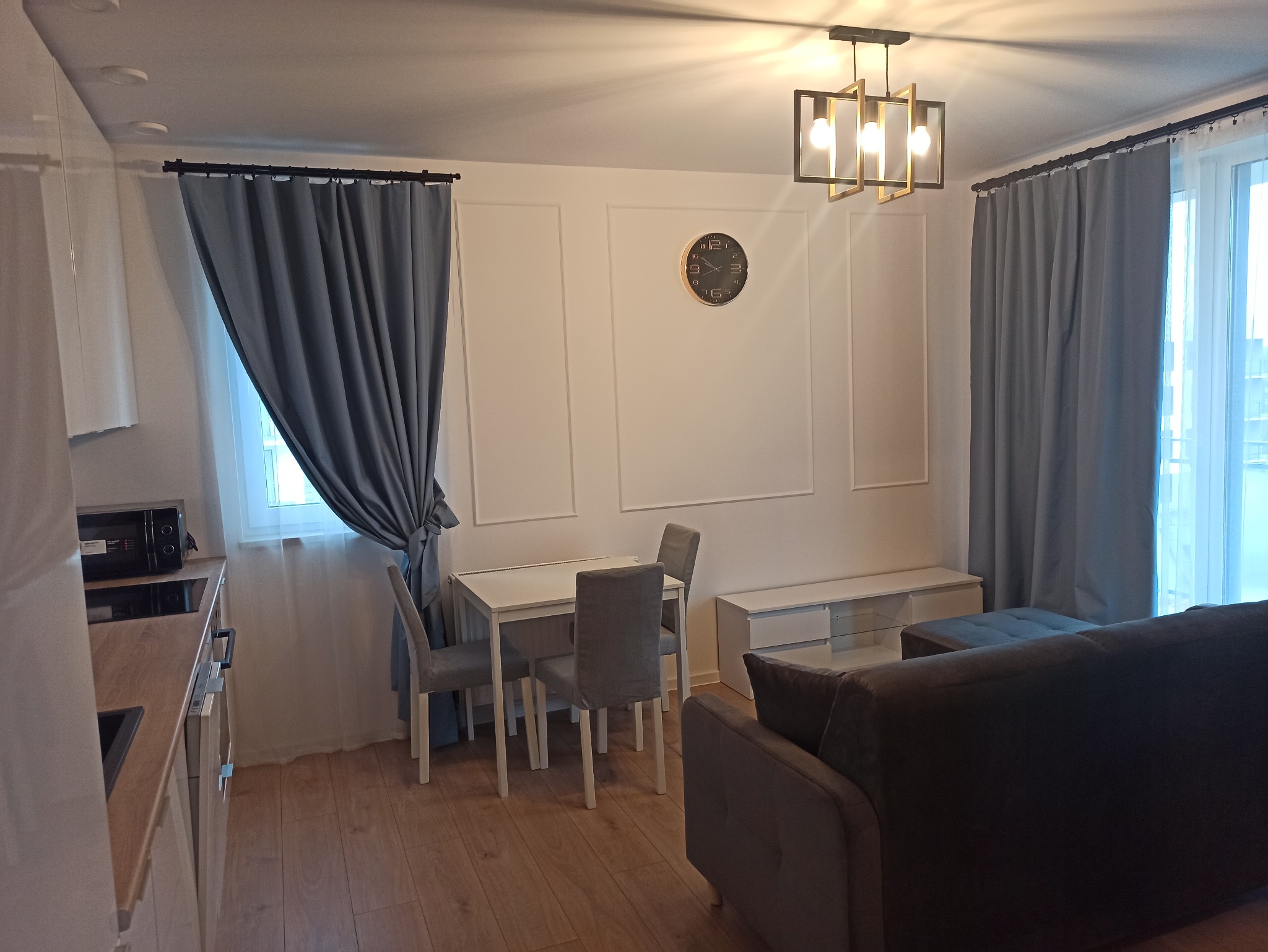 Jasne wnętrze mieszkania z szarymi zasłonami, zegarem na ścianie z dekoracyjnymi panelami, nowoczesną lampą i częściowo widoczną kuchnią.