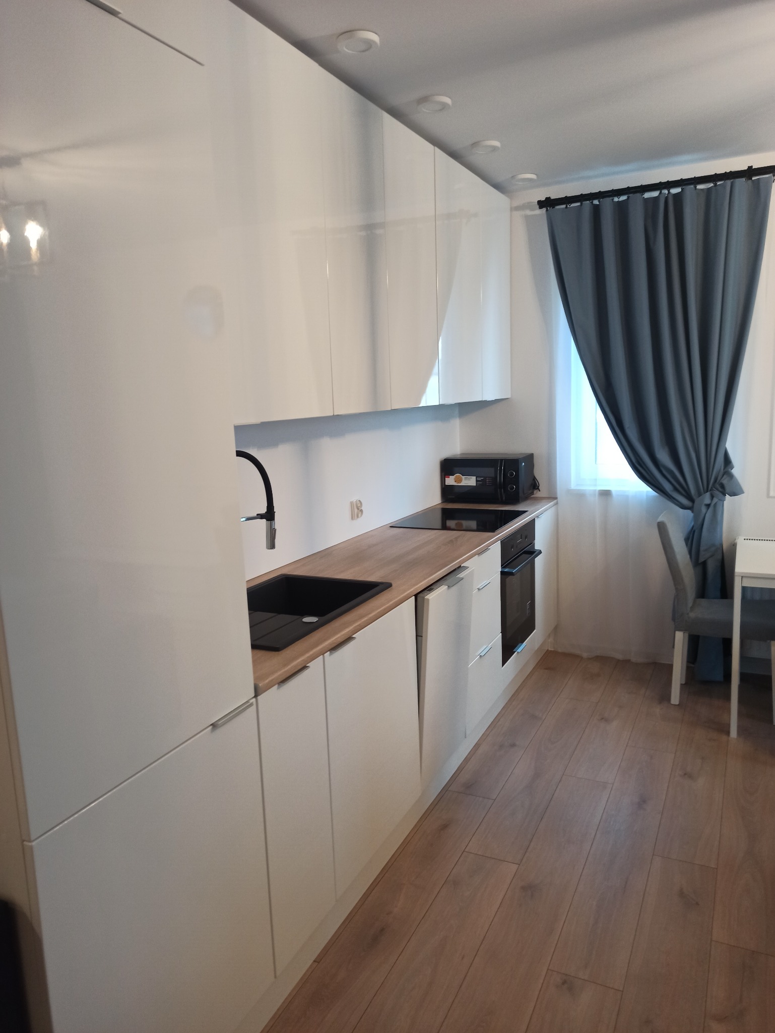 Biała, nowoczesna zabudowa kuchenna z drewnianym blatem, czarnym zlewem i baterią. W tle okno z szarą zasłoną i krzesło. Parkiet na podłodze.