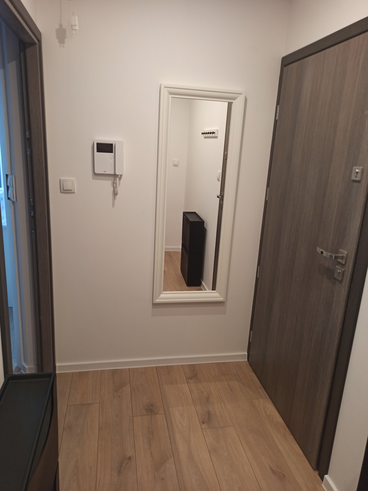 Jasne panele podłogowe w korytarzu w Gdańsku. Na ścianie wisi lustro w białej ramie, obok drzwi w odcieniach szarości i domofon. Widoczny fragment szafki na buty.