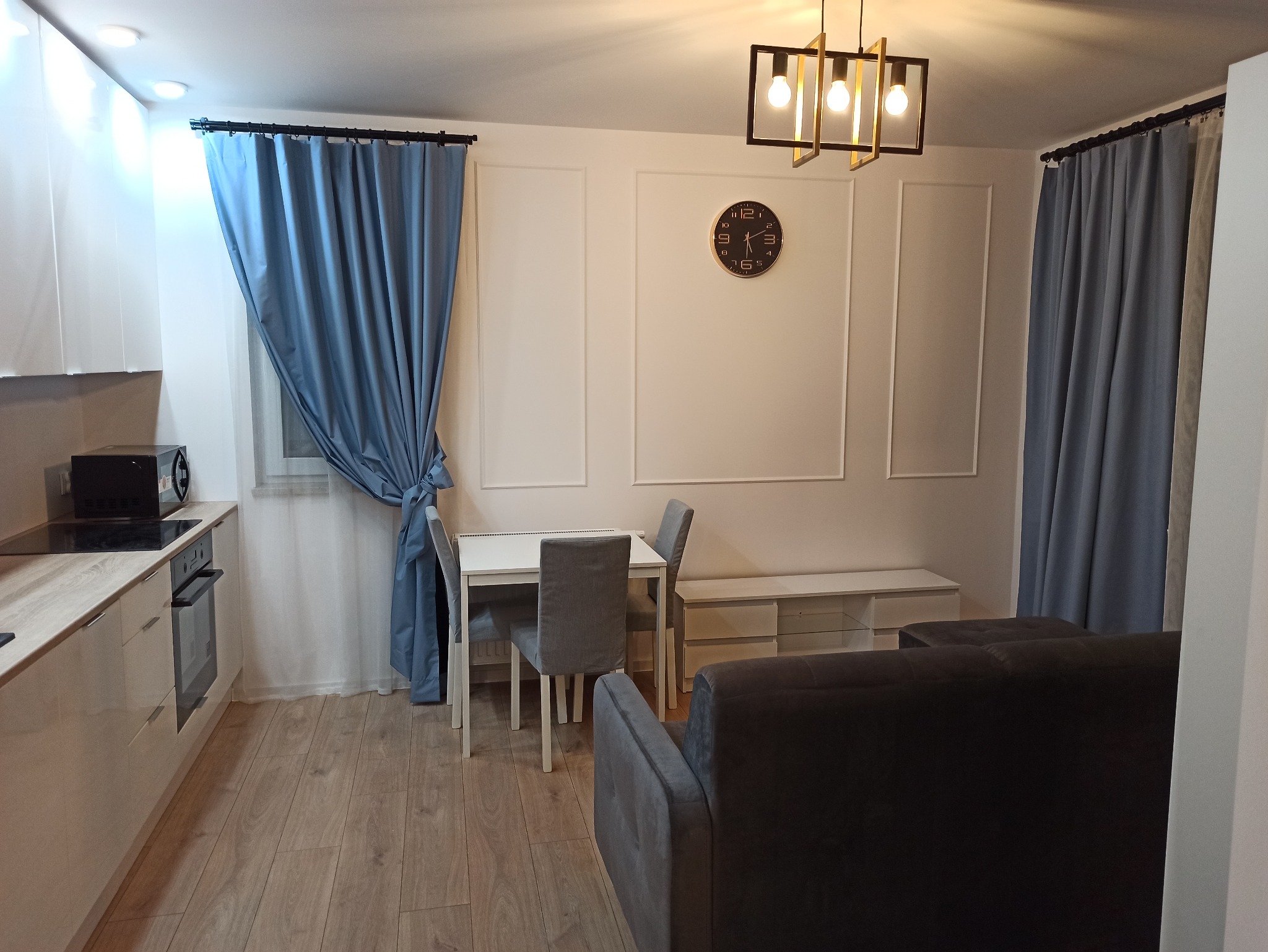 Jasne wnętrze po remoncie: kuchnia z jadalnią, drewniana podłoga, białe meble kuchenne, stół z krzesłami, szare zasłony i dekoracyjne panele ścienne.