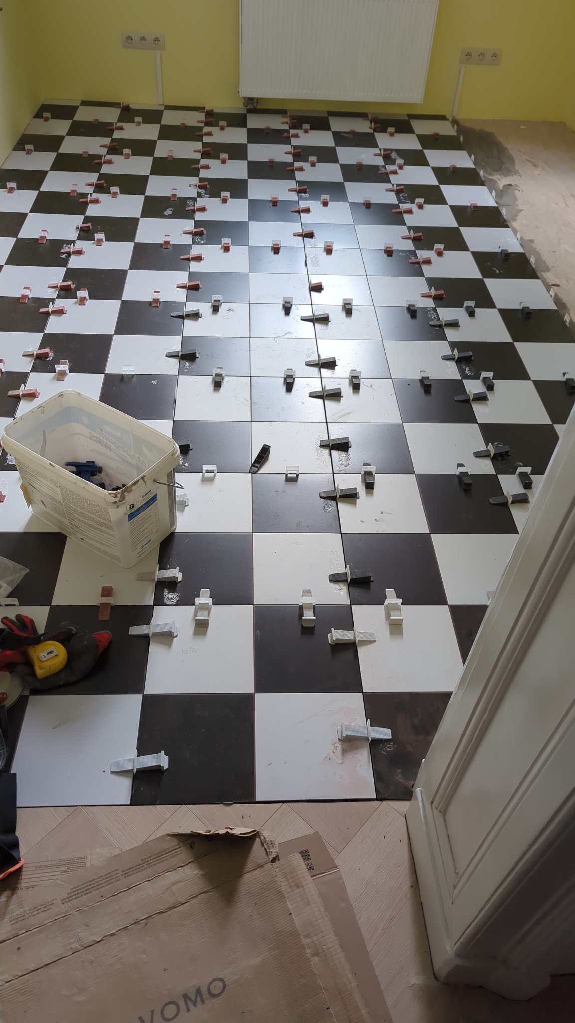 Układanie podłogi z płytek w szachownicę, z użyciem klipsów poziomujących. Widoczny pojemnik z narzędziami i fragment podłogi przed położeniem płytek.