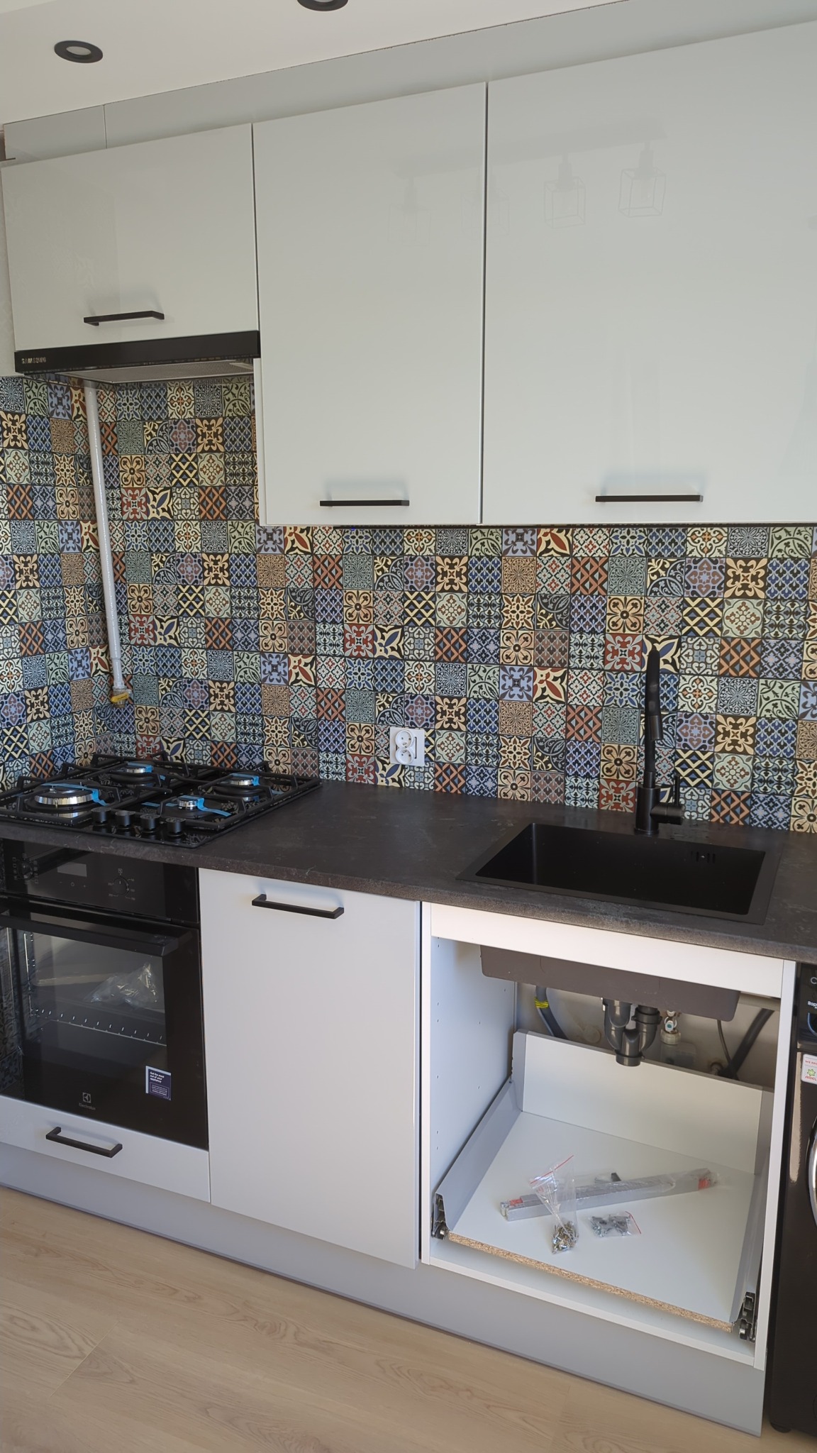 Nowoczesna kuchnia z białymi szafkami, czarnym blatem i kolorową mozaiką na ścianie. Otwarta szuflada pod zlewem z elementami montażowymi. Widoczna płyta gazowa i piekarnik.