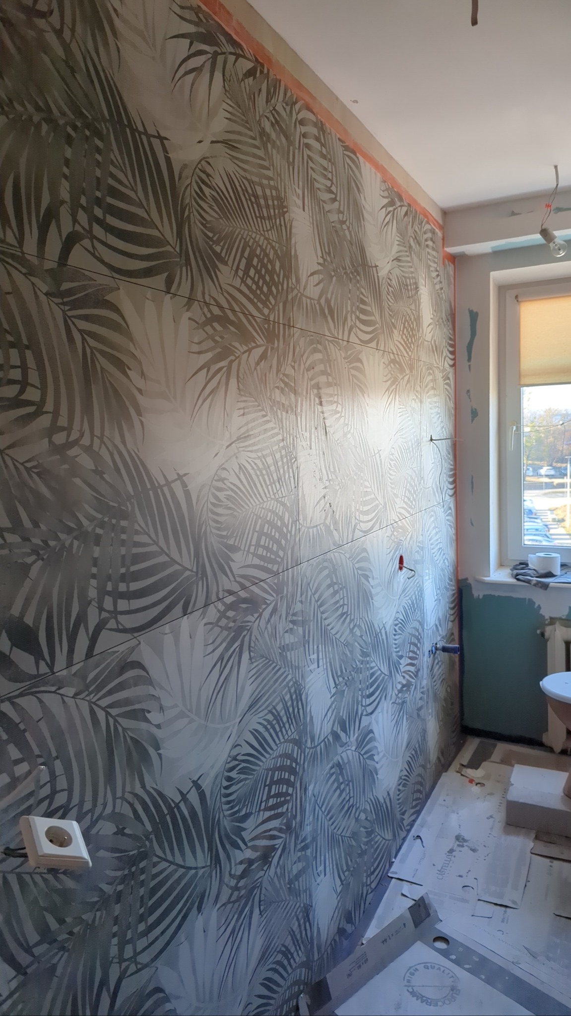 Ściana wyłożona płytkami z motywem liści palmowych w trakcie prac wykończeniowych. Widoczne narzędzia i elementy instalacji elektrycznej. Ujęcie z boku, naturalne światło.