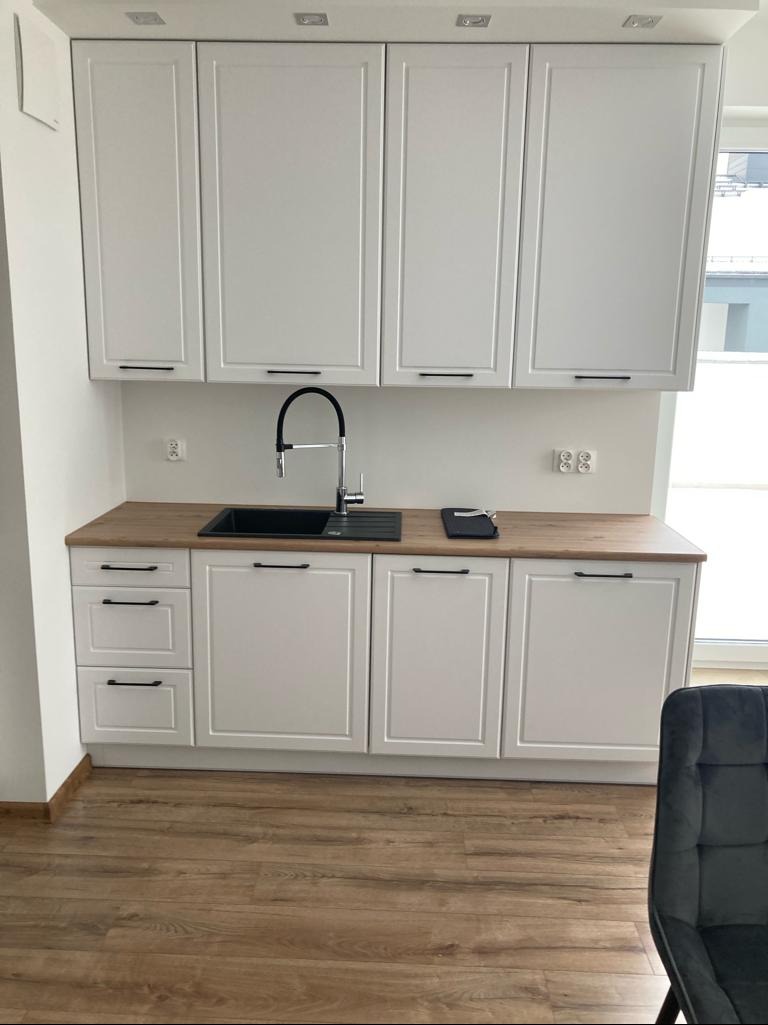 Biała zabudowa kuchenna z drewnianym blatem i czarną armaturą. Górne szafki z minimalistycznymi uchwytami. Widoczna część podłogi z paneli i fragment krzesła.