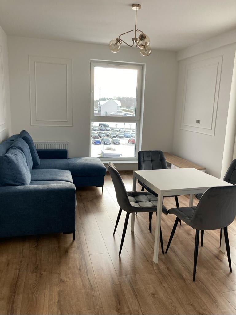 Jasne wnętrze: narożnik w odcieniach niebieskiego, stół z krzesłami na drewnianej podłodze. W tle okno z widokiem na parking i budynek. Minimalistyczny design.