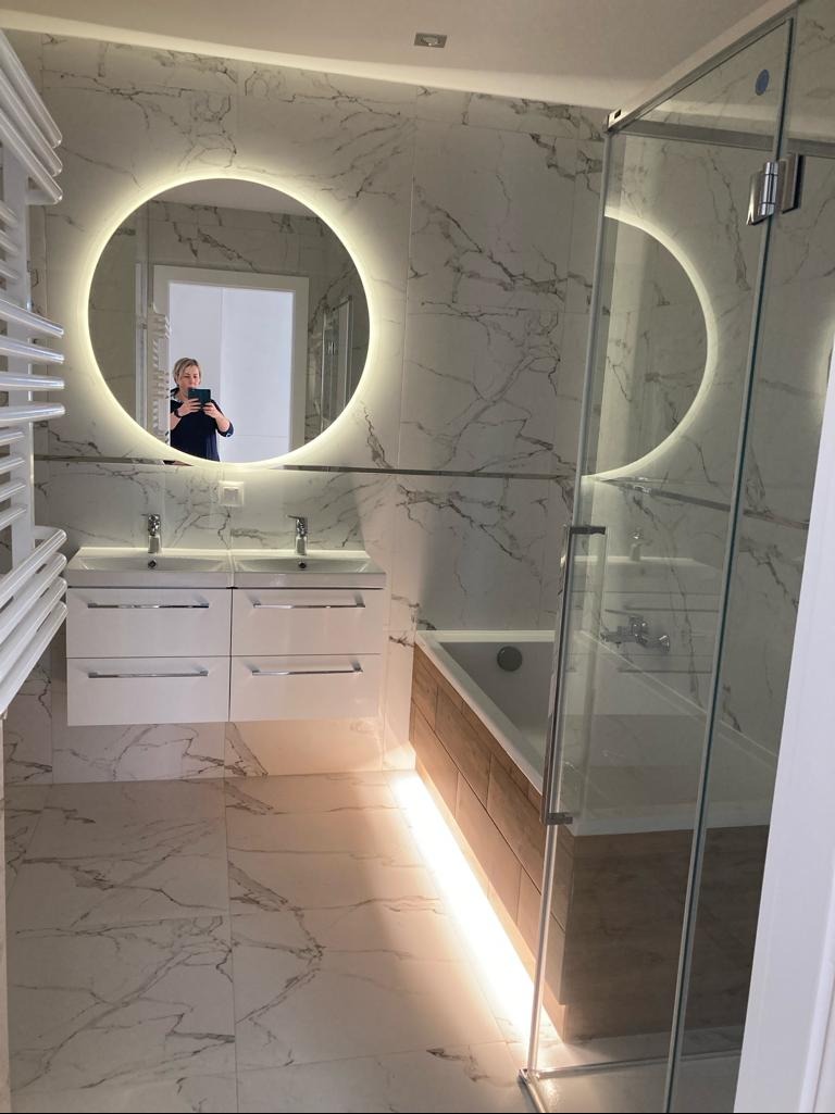Nowoczesna łazienka z marmurowymi płytkami, okrągłym lustrem z podświetleniem LED, białą szafką pod umywalką i kabiną prysznicową ze szklanymi drzwiami.