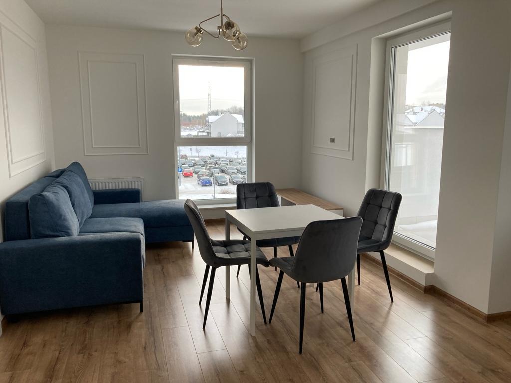 Jasne wnętrze salonu z niebieską sofą narożną, białym stołem i szarymi krzesłami. Widok na parking przez okno. Parkiet na podłodze.