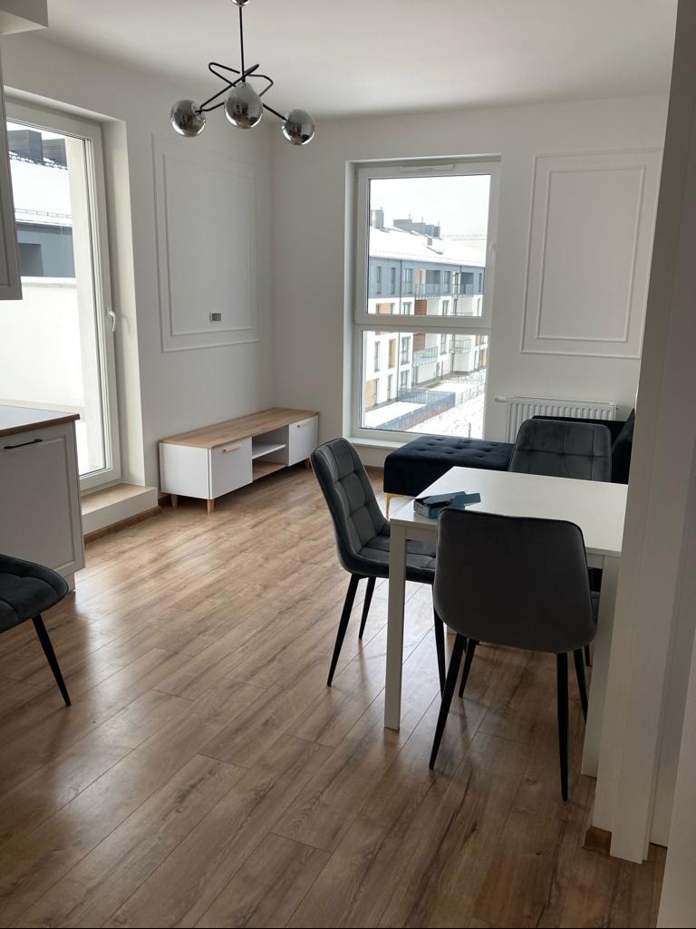 Jasne wnętrze pokoju dziennego z drewnianą podłogą, białym stołem i szarymi krzesłami. W tle okno z widokiem na budynki mieszkalne i dekoracyjne listwy na ścianach.