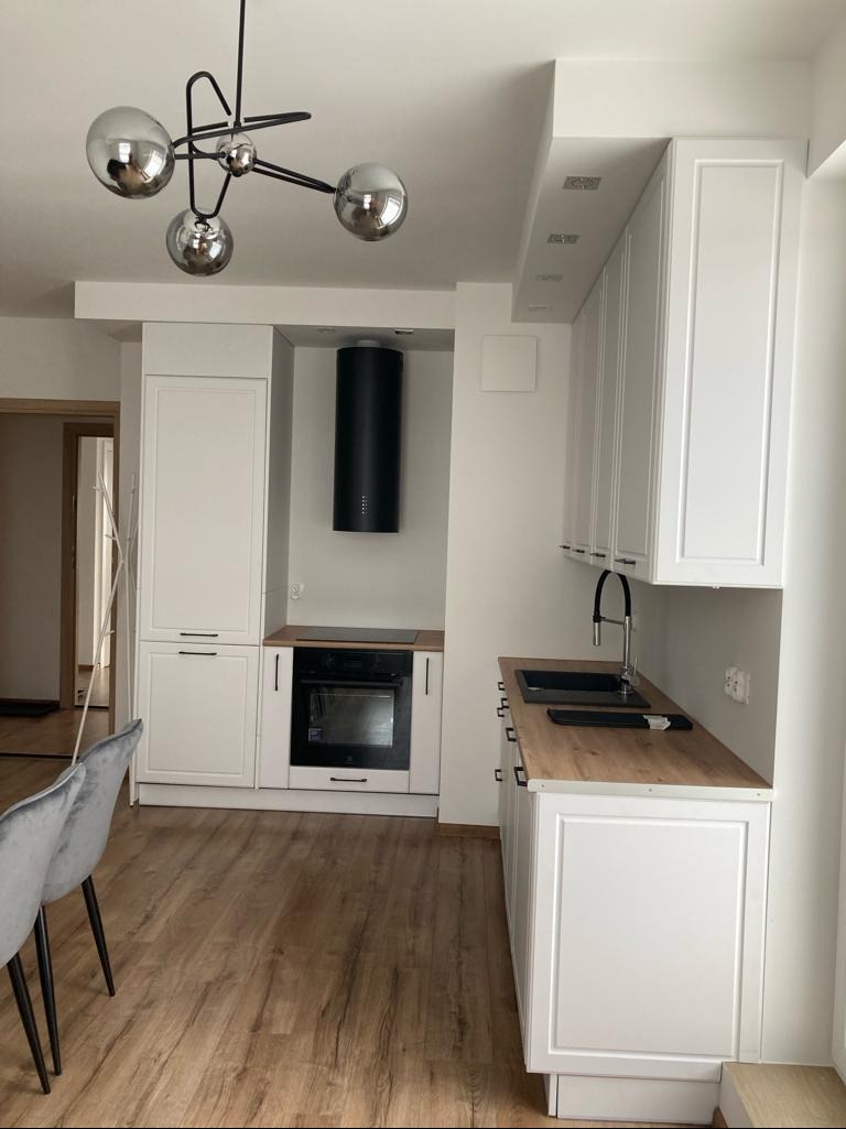 Biała zabudowa kuchenna z drewnianym blatem i nowoczesną lampą w Gdańsku. Minimalistyczny design, czarny okap i jasna podłoga.
