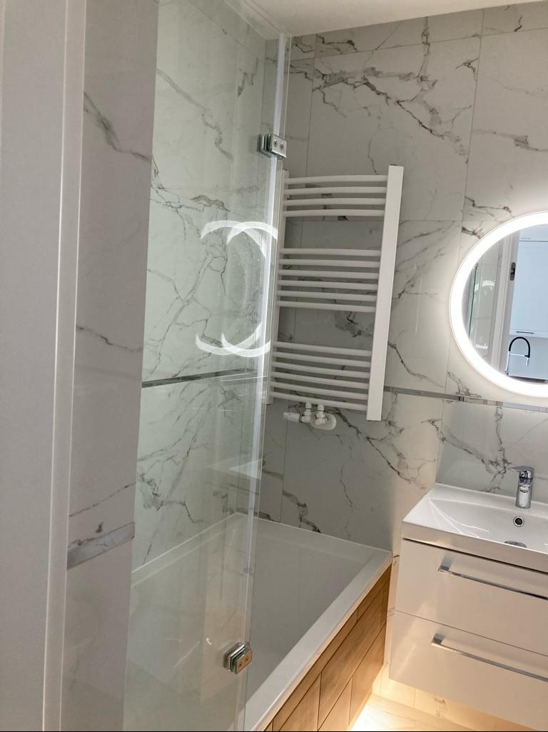 Nowoczesna łazienka z kabiną prysznicową ze szklanymi drzwiami, grzejnikiem drabinkowym i umywalką z szafką. Marmurowe płytki na ścianach i oświetlone lustro.