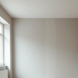 Krzysztof Wojciechowski - Jasne wnętrze po remoncie: ściany pomalowane na szaro, jedna ściana z dekoracyjną fakturą, białe okno z grzejnikiem pod spodem. Minimalistyczny design.