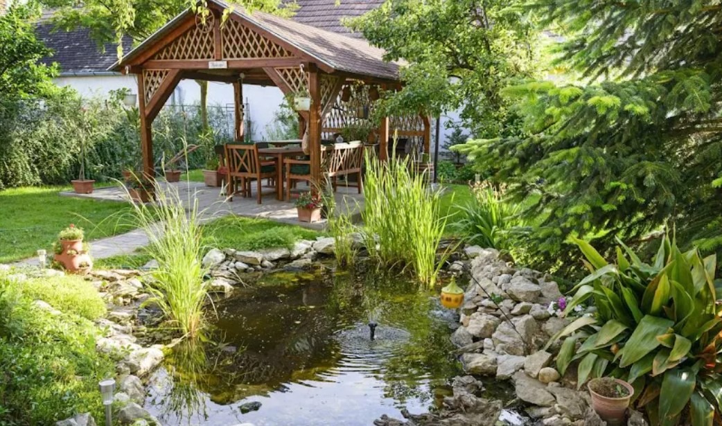 Ogród z oczkiem wodnym i altaną z drewnianą konstrukcją. Woda odbija zieleń, a kamienie tworzą naturalny brzeg. W tle widoczny dom i drzewa.