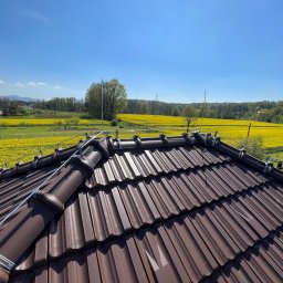 Instalatorstwo elektryczne Herzyk Łukasz - Brunatny dach z dachówki ceramicznej z zamontowaną instalacją odgromową na tle kwitnącego pola rzepaku i błękitnego nieba w słoneczny dzień.