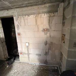 Instalatorstwo elektryczne Herzyk Łukasz - Surowy stan: instalacja elektryczna w trakcie montażu w pomieszczeniu z betonu komórkowego. Widoczne kable, puszki instalacyjne i rury. Podłoże nieutwardzone.