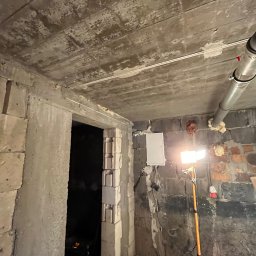 Instalatorstwo elektryczne Herzyk Łukasz - Surowy stan: biała skrzynka elektryczna na ceglanej ścianie, oświetlona lampą budowlaną. Widoczne kable i rury. W tle surowy betonowy strop i otwór drzwiowy.