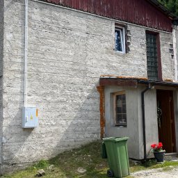 Instalatorstwo elektryczne Herzyk Łukasz - Szara skrzynka elektryczna z białymi rurami na szarej, chropowatej ścianie budynku z czerwonymi elementami drewnianymi. Widoczny fragment trawnika i ścieżki.