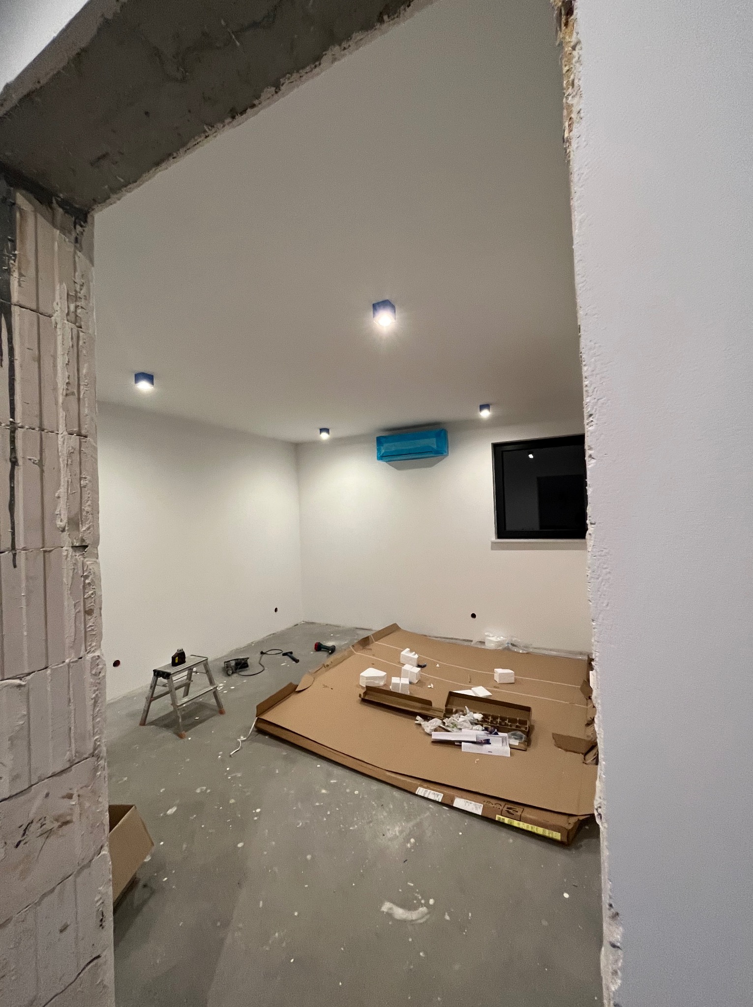 Wnętrze w trakcie wykańczania: widoczne punkty oświetleniowe w suficie, klimatyzator zabezpieczony folią, narzędzia i materiały na podłodze. Surowy stan wykończenia.