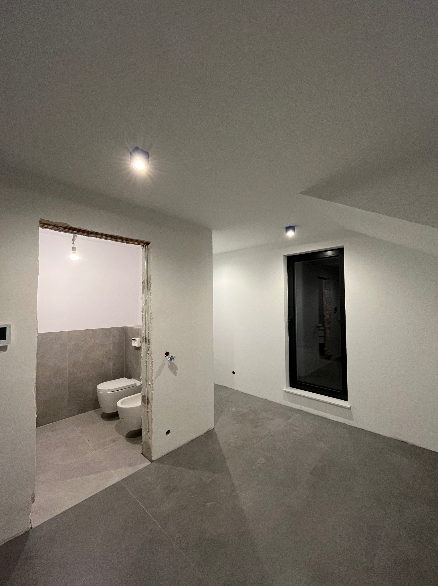 Wnętrze w trakcie remontu: szara podłoga, białe ściany, czarne okno. Widoczne przygotowane wejście do łazienki z zamontowaną armaturą. Dwa punkty oświetleniowe na suficie.