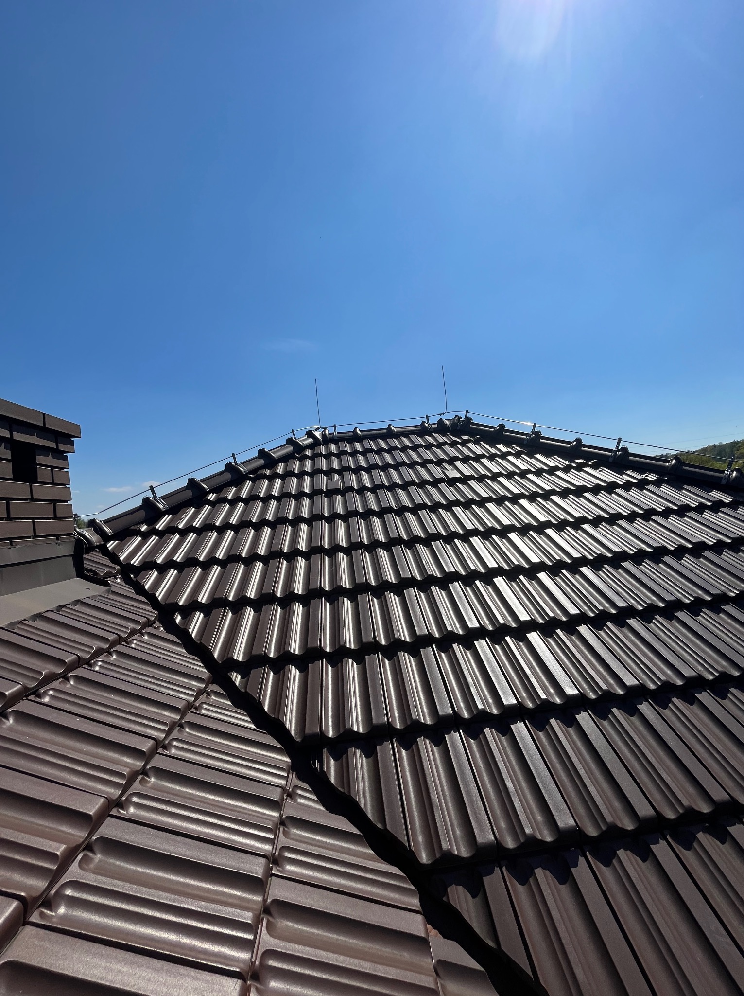 Błyszczący, brązowy dach z dachówki ceramicznej, zabezpieczony odgromowo, na tle błękitnego nieba i komina z cegły klinkierowej. Widok z góry.