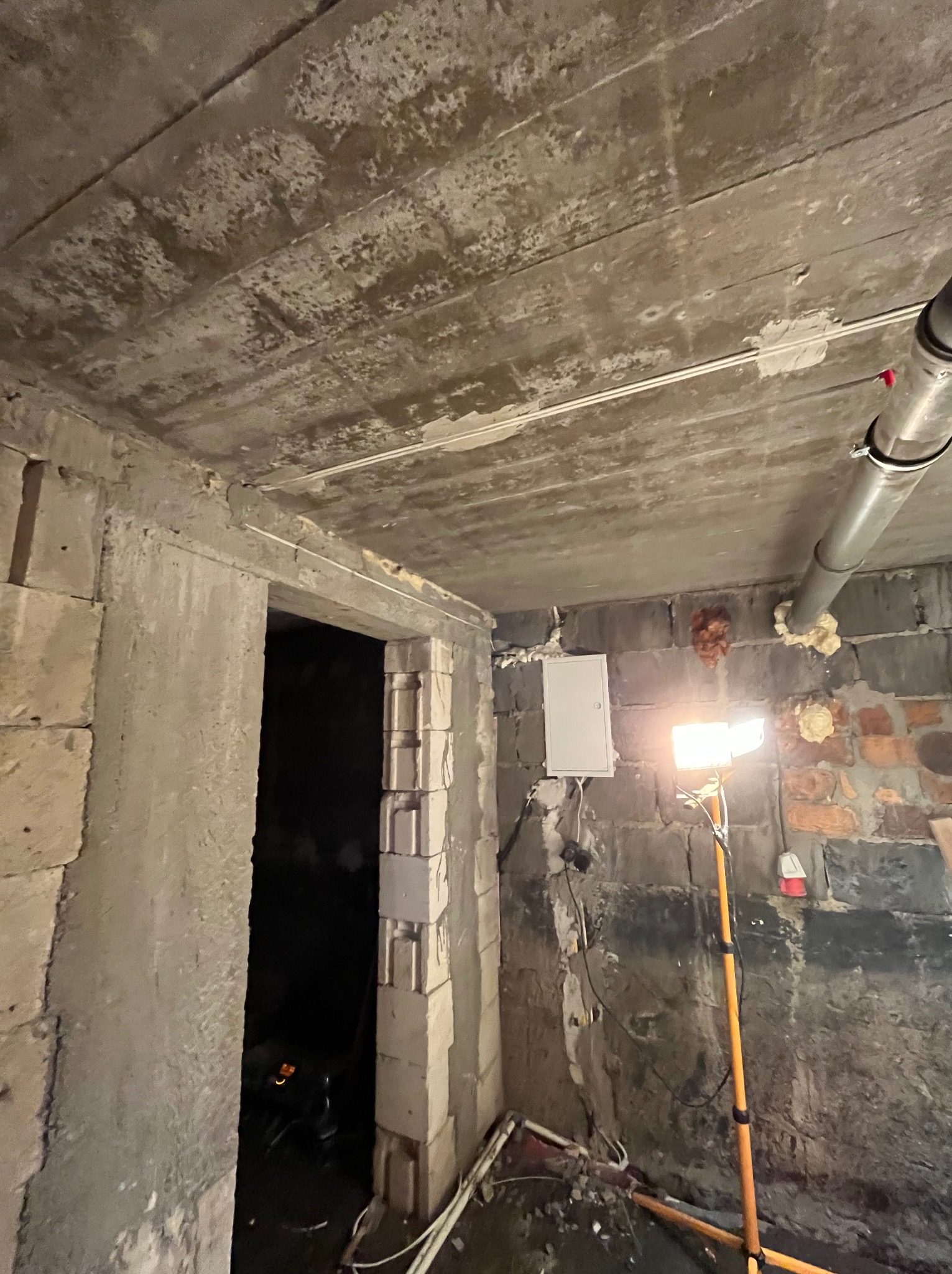 Surowy stan: biała skrzynka elektryczna na ceglanej ścianie, oświetlona lampą budowlaną. Widoczne kable i rury. W tle surowy betonowy strop i otwór drzwiowy.